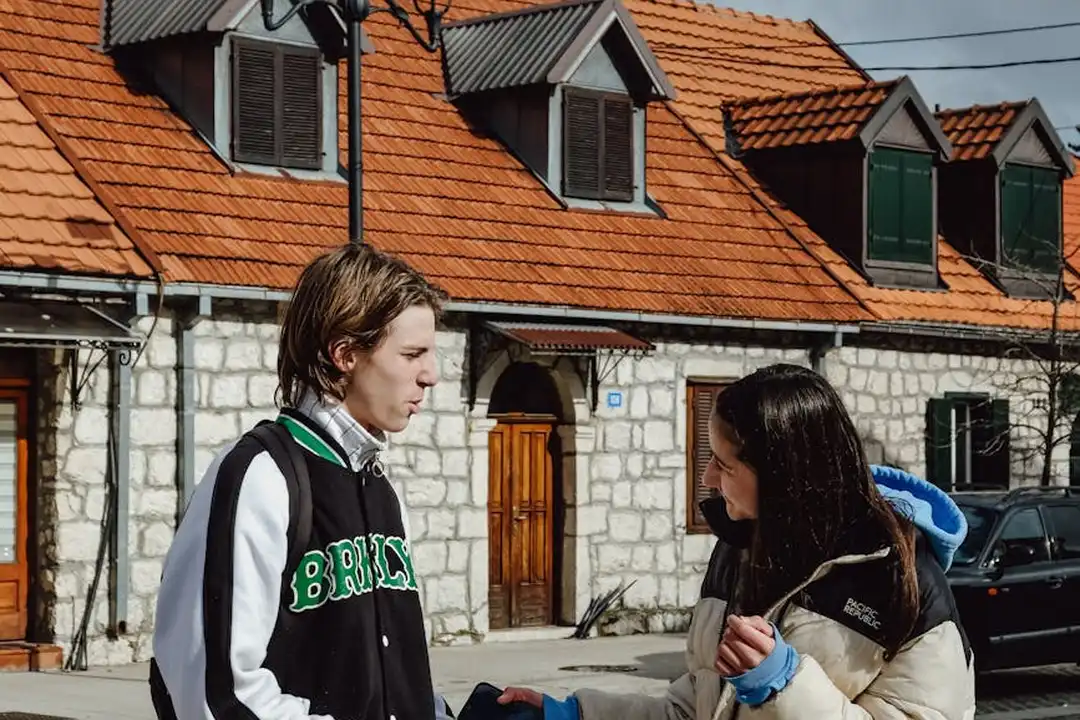 Two neighbors standing and talking outdoors in front of a row of brick houses with orange-tiled roofs, illustrating stakeholder conversations for HOA communication planning.