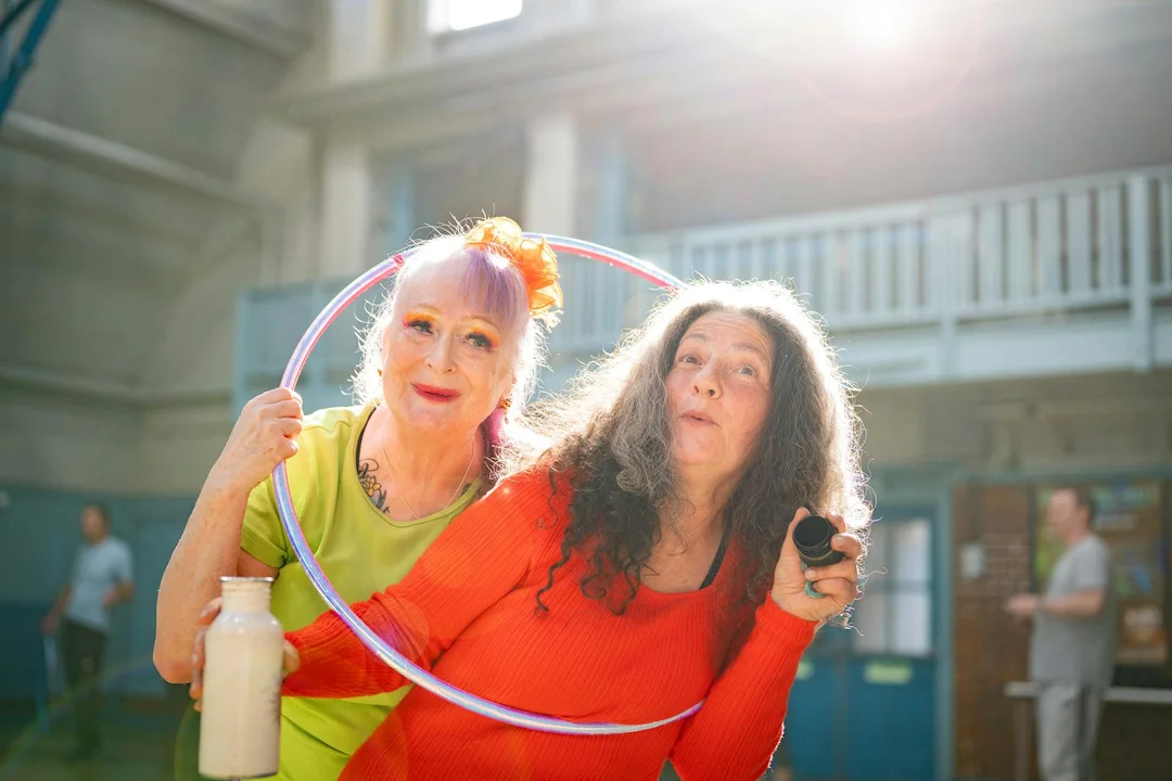 Two women at an HOA community gym pose with a hula hoop; one wears a lime green shirt and a flower in her hair, the other wears a red shirt and holds a small device, with a water bottle visible nearby.