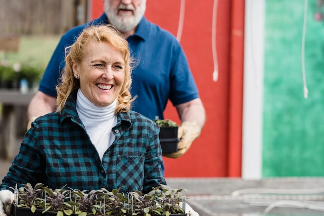 Smiling woman in a plaid shirt holds a tray of young seedlings in a community garden while a man in the background tends plants.