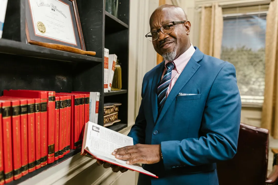 A professional man in a blue suit stands beside a bookshelf with legal certificates, holding an open book in a well-lit office.