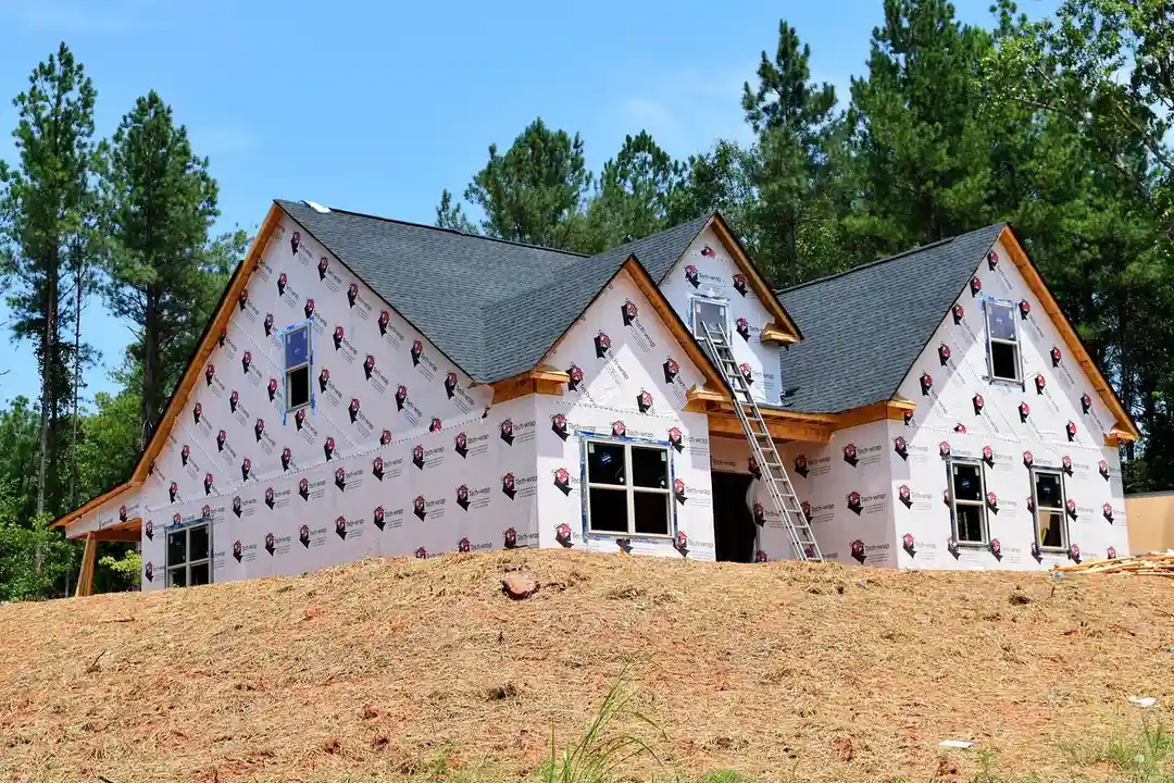 Residential construction site with an unfinished house wrapped in protective sheathing, a ladder leaning against the front, and trees in the background.