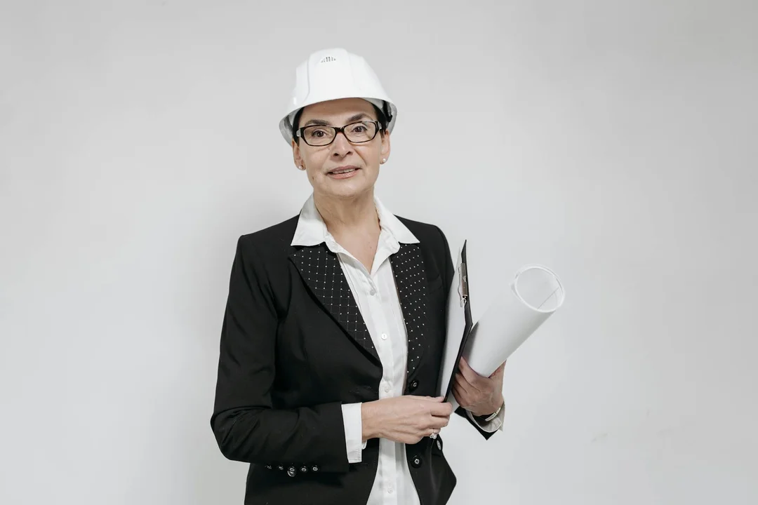 Professional woman wearing a white hard hat and blazer, holding rolled blueprints.