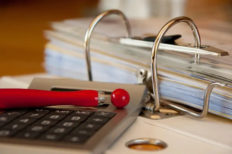 Close-up of a binder with stacked documents, a calculator, and a red pen on a desk, illustrating reviewing HOA maintenance decisions and fees.