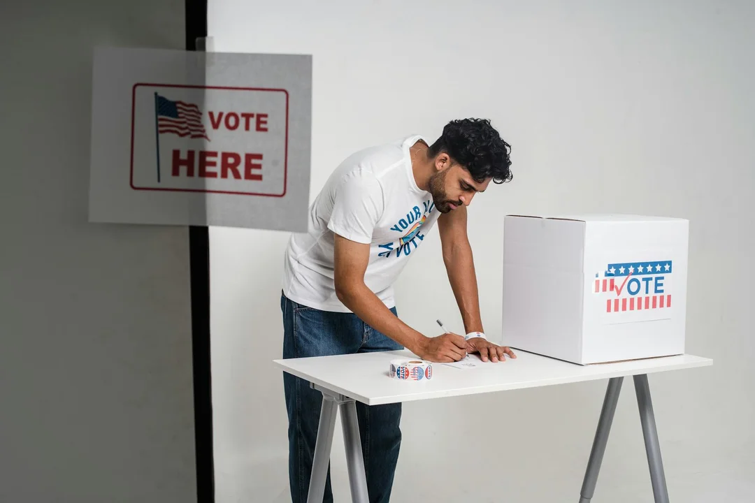 Man signing a ballot at a table with a white voting box labeled 'I VOTE', beside a wall sign reading 'VOTE HERE'.
