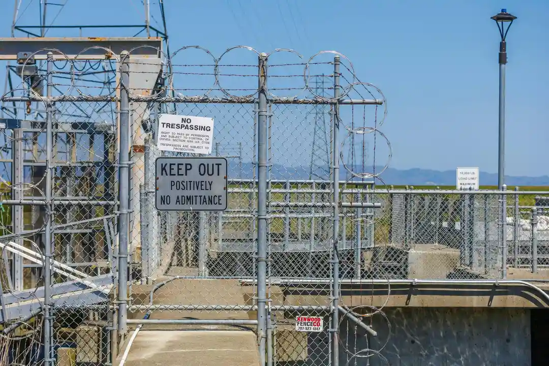 Secure chain-link fence with barbed wire and multiple warning signs reading 'Keep Out' and 'No Trespassing,' surrounding a gated facility near a body of water.