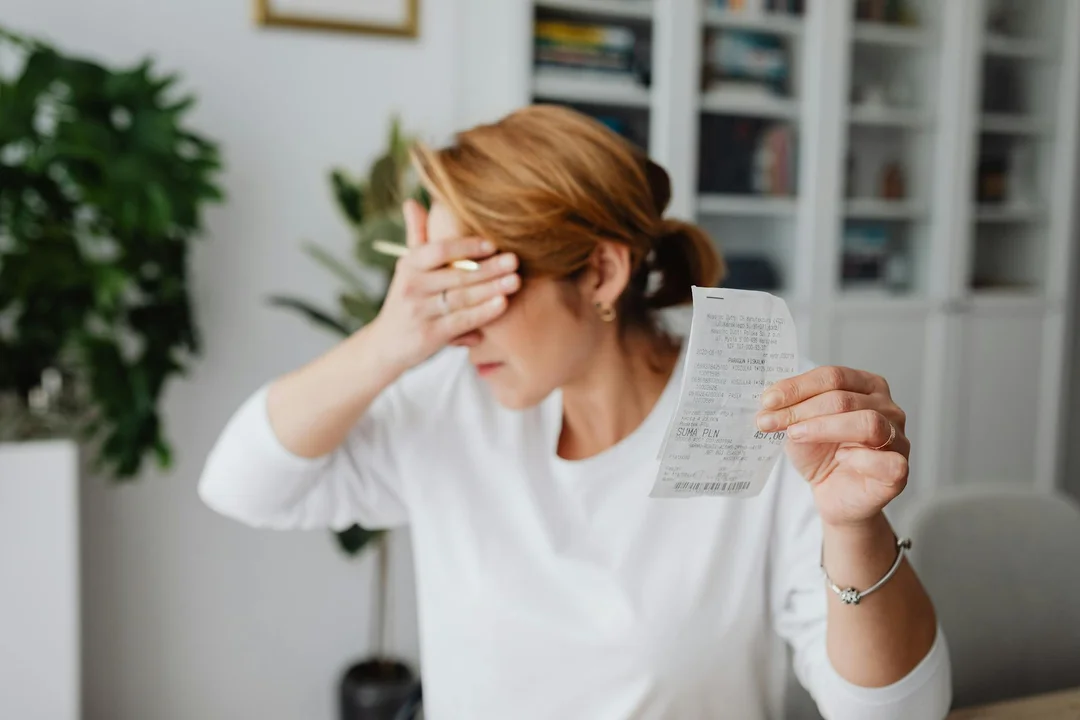 A stressed-looking woman sits at a table, holding a receipt in one hand and shielding her eyes with the other, illustrating concerns about HOA payment timing.