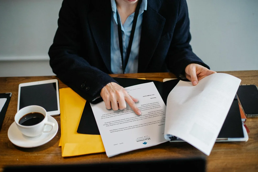 A business professional in a dark blazer sits at a wooden desk, flipping through documents with a coffee cup, tablet, and yellow folder nearby.