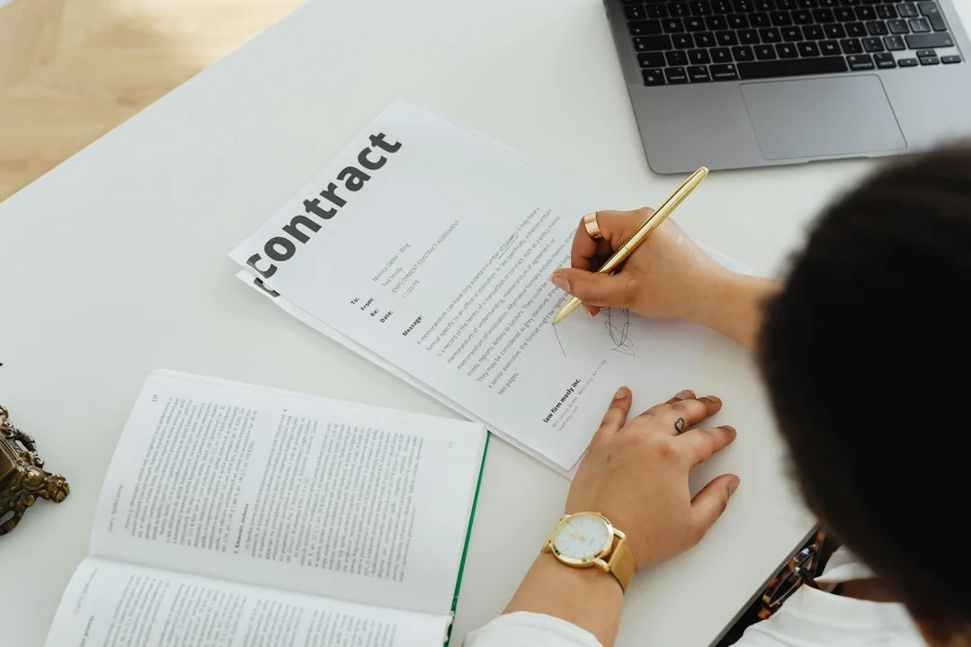 Person's hands at a desk reviewing a contract and an open book, with a laptop nearby, symbolizing financial oversight and governance.