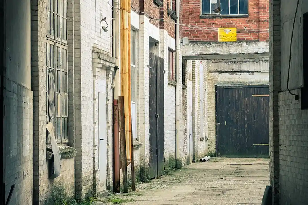 Narrow urban alley with weathered brick buildings, closed doors, and a dark garage door at the end.