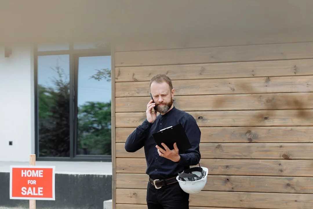 Man in a dark shirt stands on a construction site outside a home, talking on his phone while holding a clipboard with a white hard hat tucked under his arm, near a 'Home For Sale' sign.