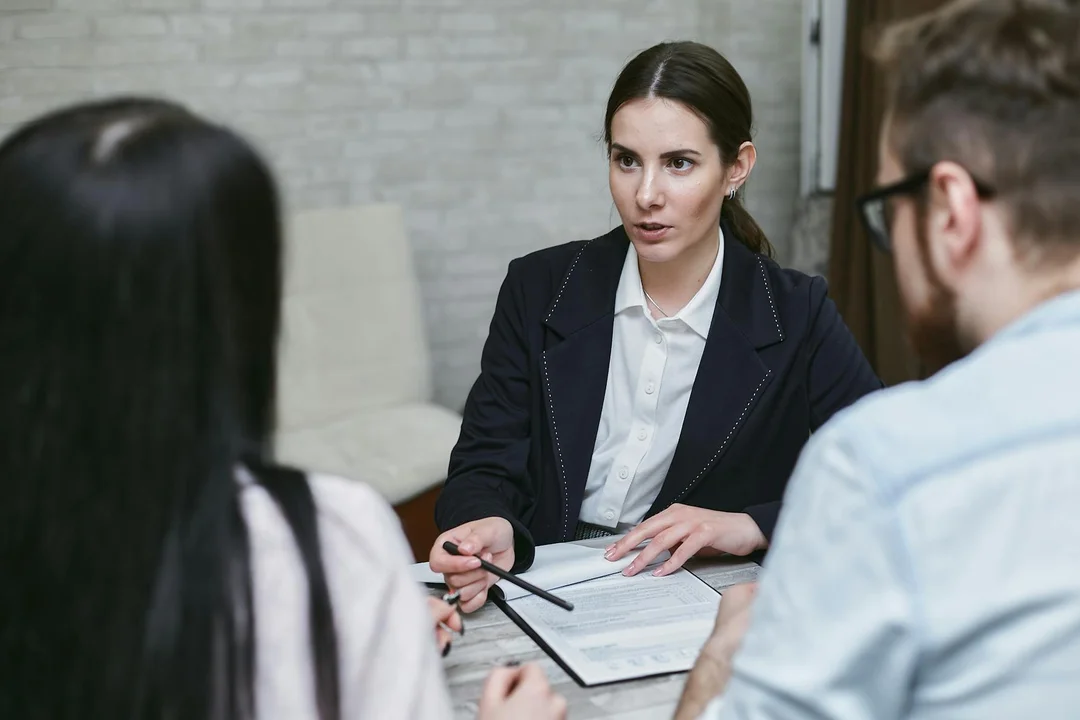 A business meeting with a woman in a dark blazer presenting documents to two people seated across a table, emphasizing legal and financial safeguards during a developer handoff.
