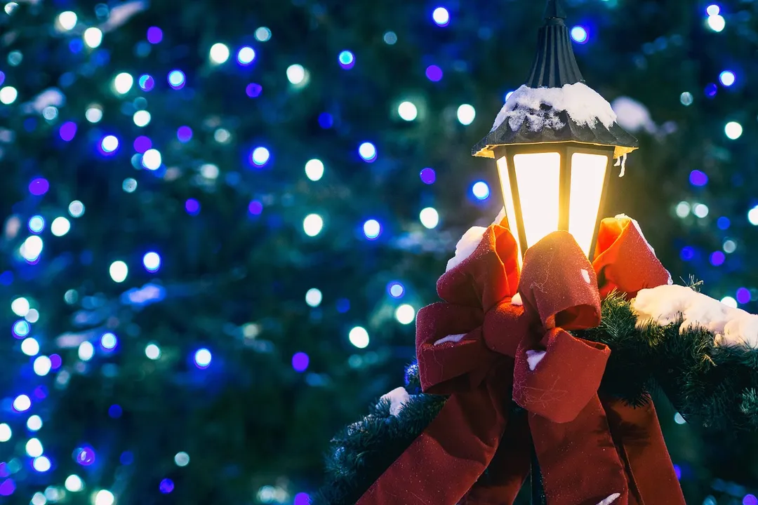 Snow-dusted lamppost adorned with a red ribbon and warm glow, with blue holiday lights softly blurred in the background.
