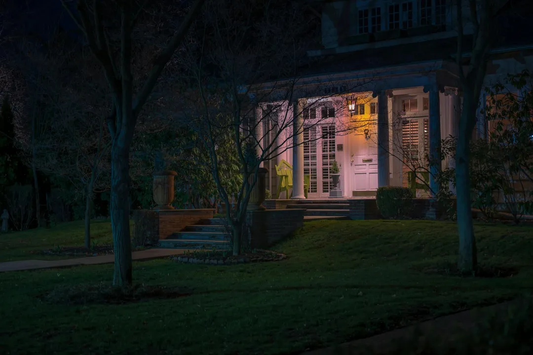 Nighttime view of a well-lit front porch of a house in an HOA community, with a manicured lawn and surrounding trees.