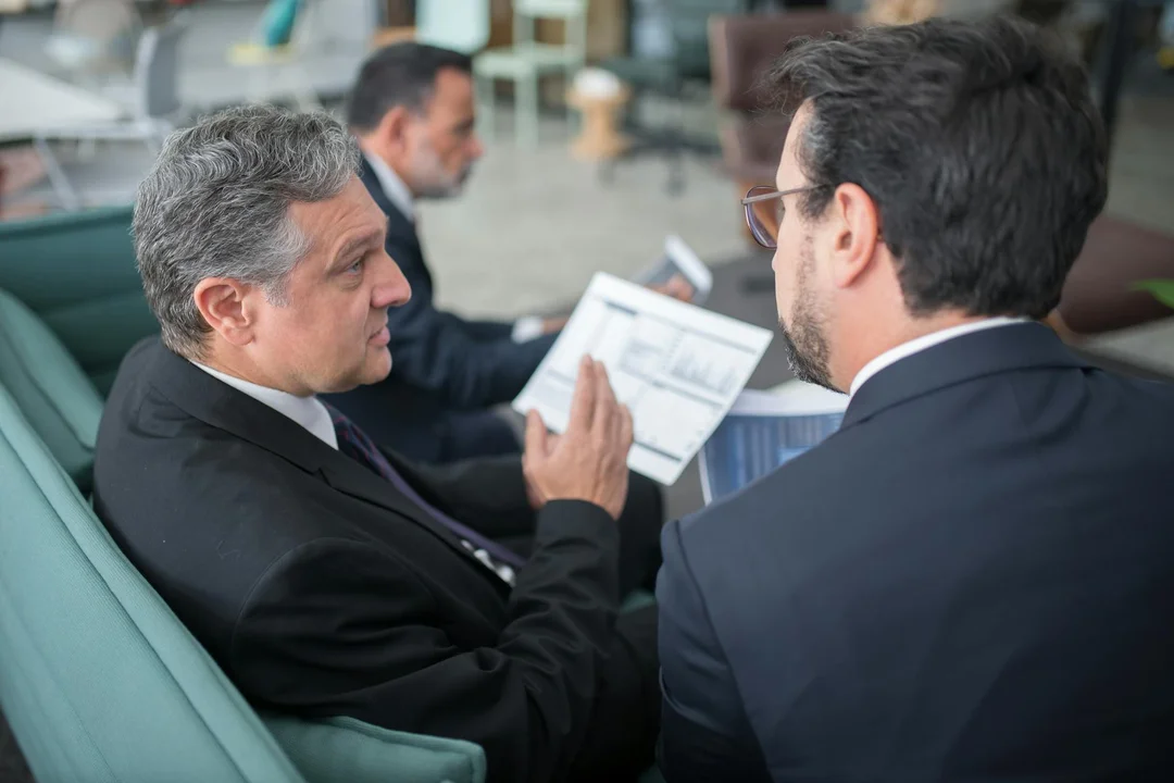 Two men in business suits sit in a modern lobby, facing each other and reviewing a document together.