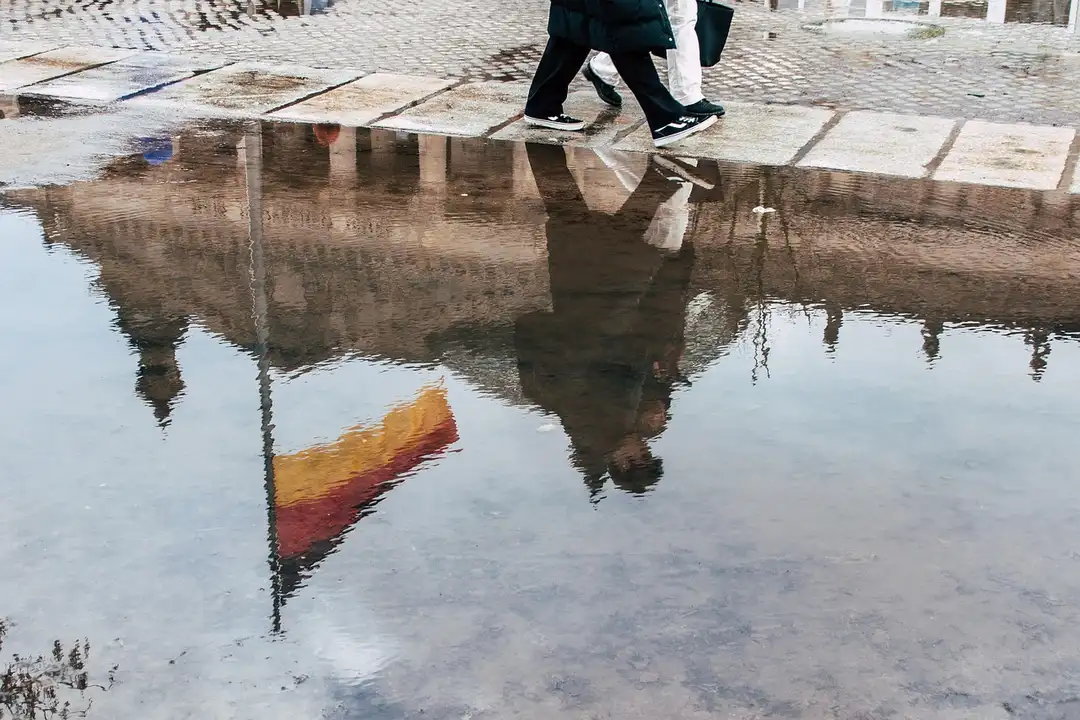 Two people walk along a wet sidewalk with a large puddle, reflections of surrounding buildings and street visible in the water.