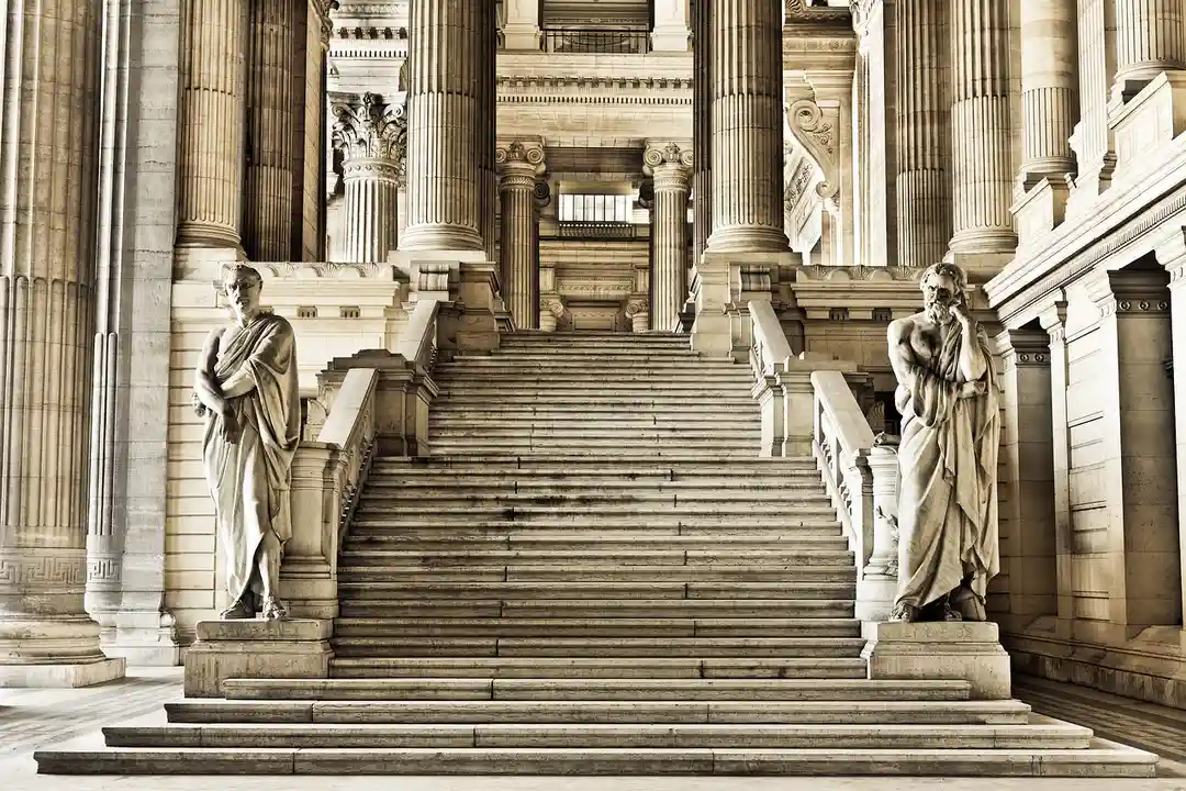 Two stone statues flank a grand staircase in a neoclassical building with tall columns and ornate architecture.