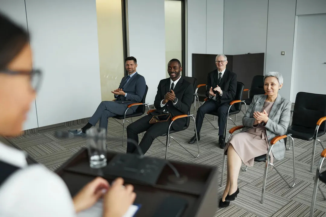 A presenter at a podium addresses a small audience of four professionals seated in a modern conference room.