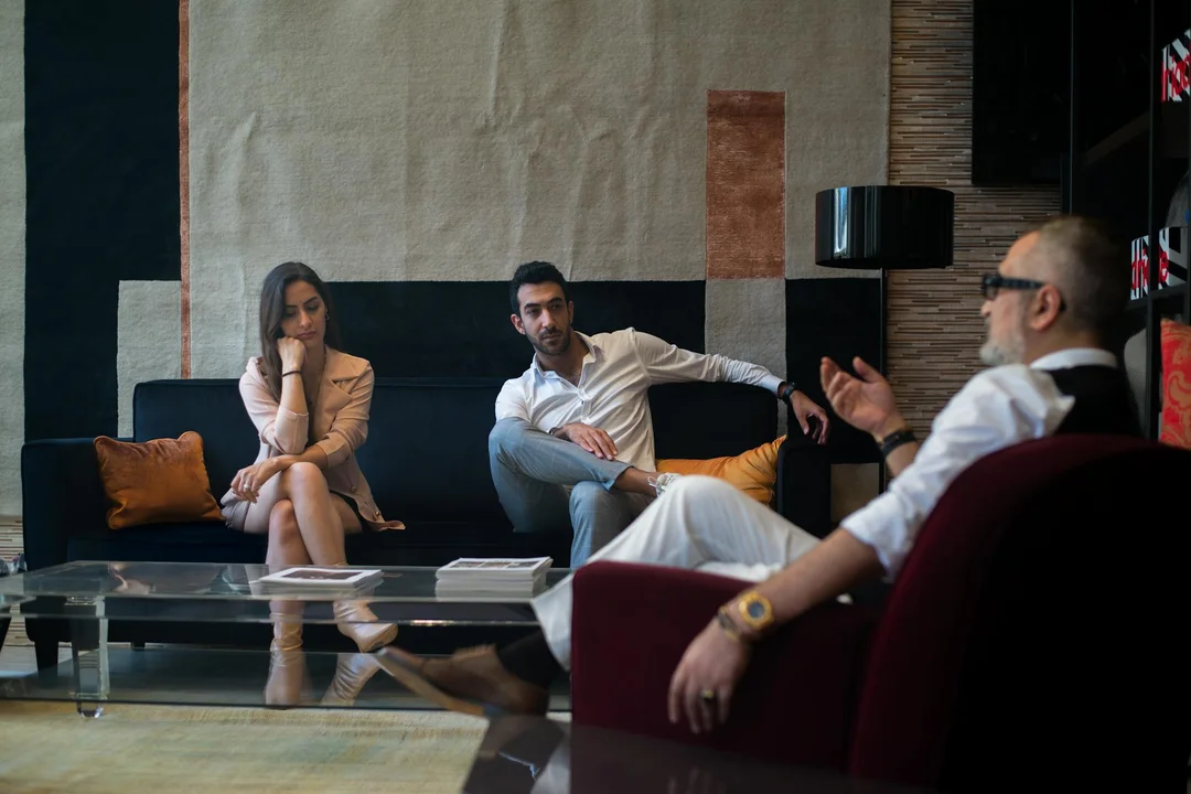 Three adults in a modern living room sit around a glass coffee table with documents, engaging in a mediation discussion about HOA disputes.