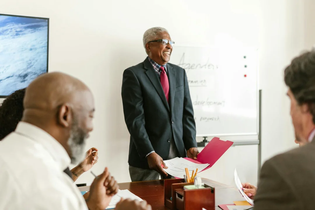 An HOA meeting facilitator stands at the head of a conference table presenting to residents, with a whiteboard in the background and documents on the table.