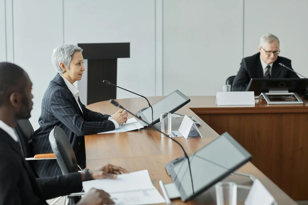 A formal HOA board meeting in a conference room with a woman speaking into a microphone at the table while two other attendees listen.
