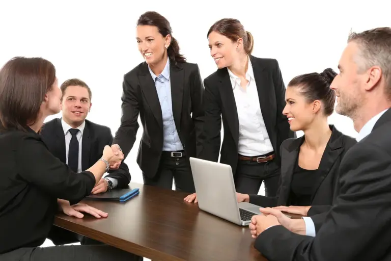 Group of professionals in business attire seated around a conference table; two people are shaking hands while others watch and a laptop sits on the table.