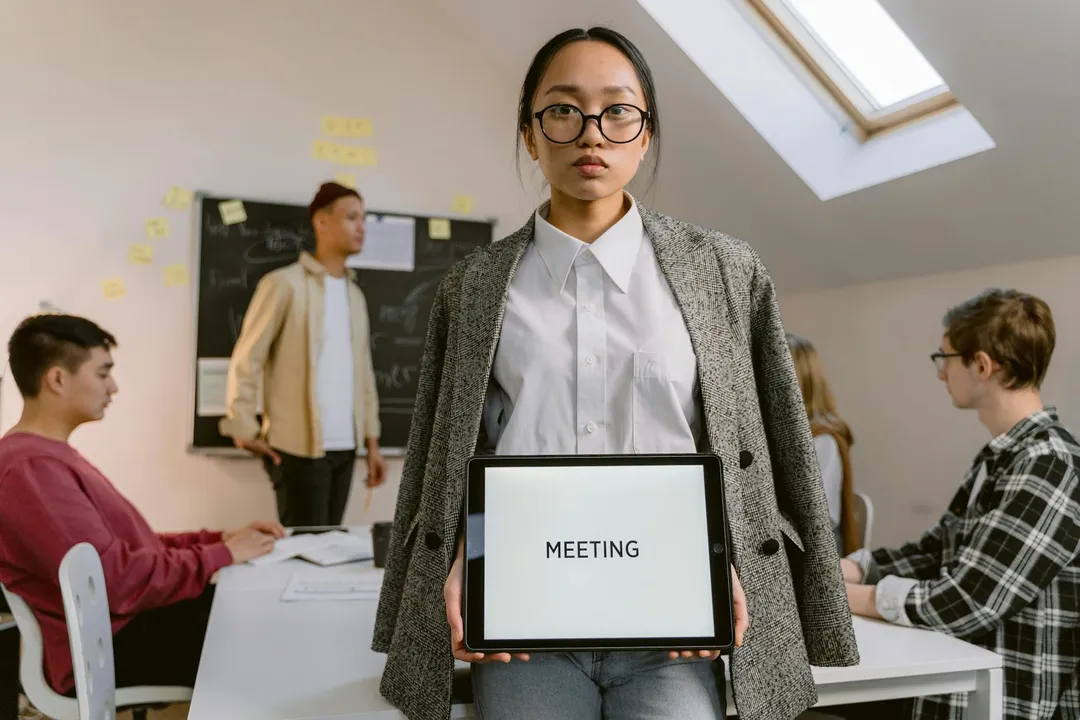 A woman wearing glasses and a blazer holds a sign reading 'MEETING' in a conference room, with other residents seated and discussing in the background.