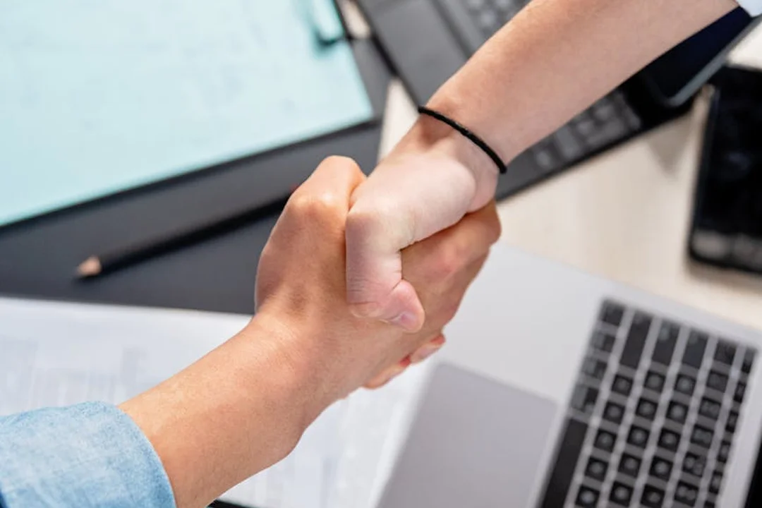 Two hands shake over a desk with a laptop and documents, representing direct contact at HOA meetings.