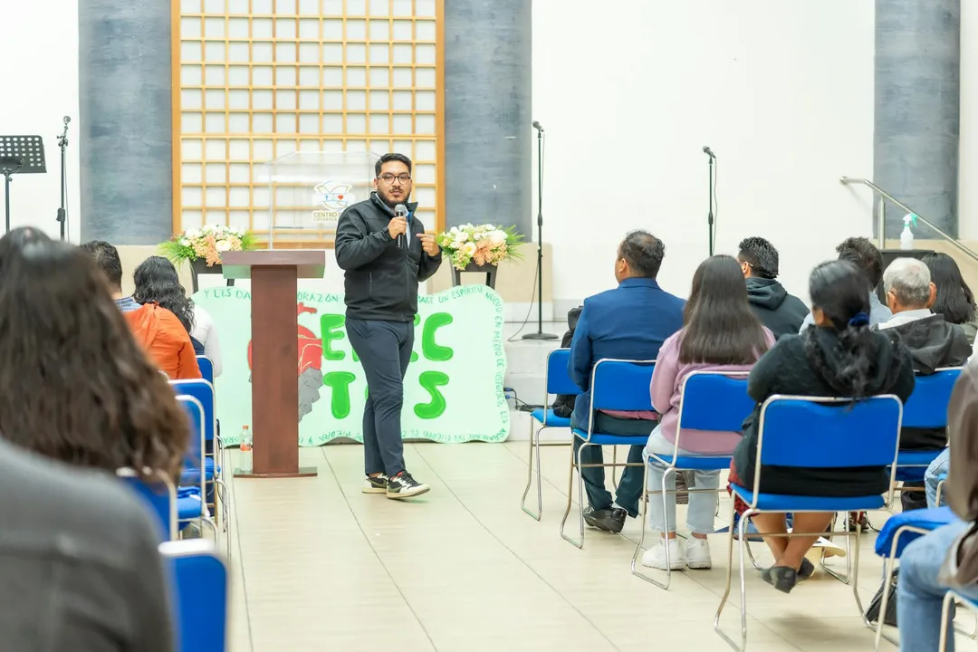 Speaker at a podium addressing homeowners in an HOA meeting; audience seated in blue chairs in a bright room, with a banner and flowers visible in the background.