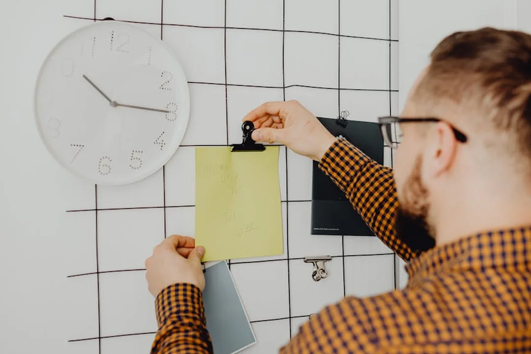 A person in a checkered shirt pins a yellow sticky note to a grid wall beside a large white clock, illustrating planning for post-meeting follow-up tasks.