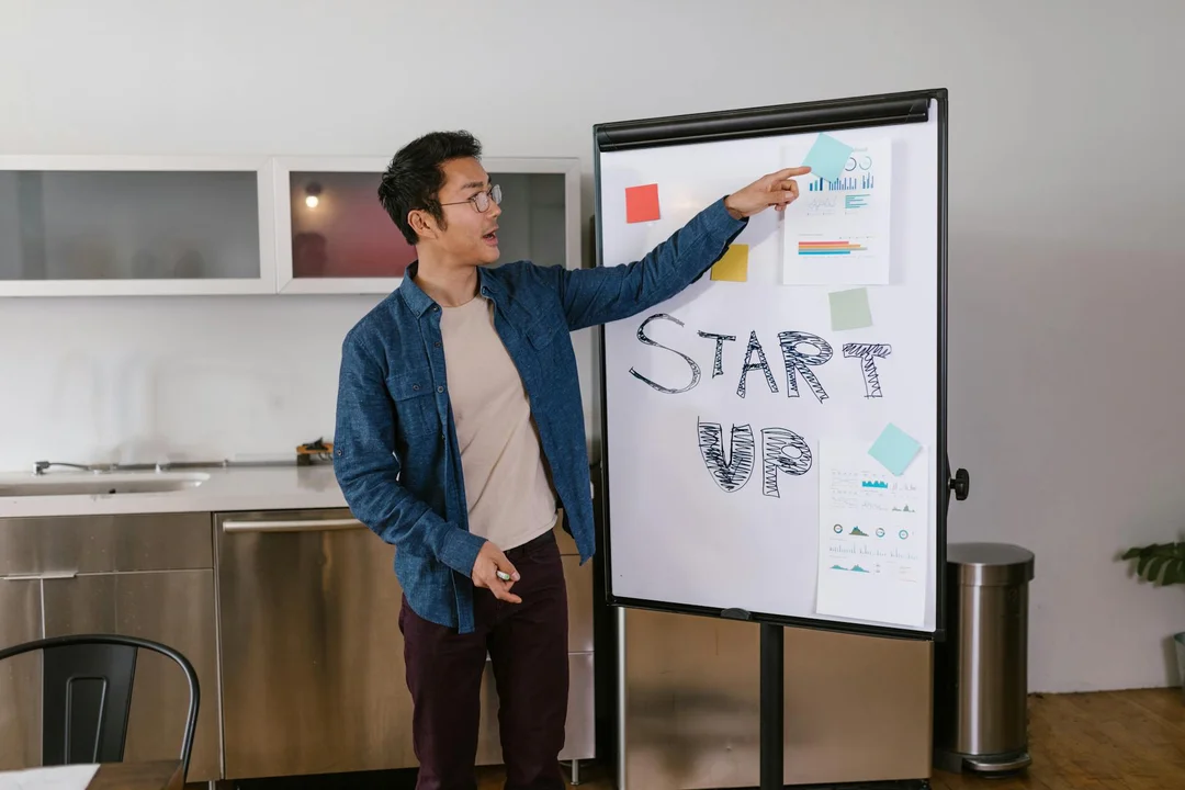 A man in a denim jacket points to a flip chart with charts and the word 'START VP' in a modern meeting room.