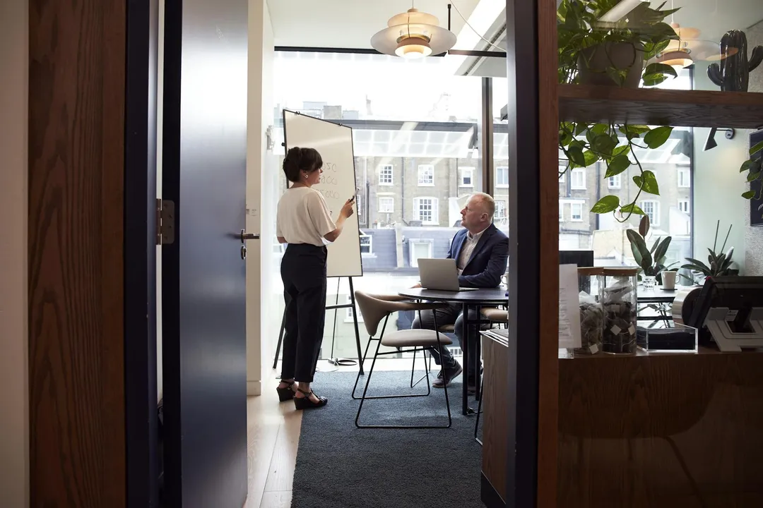A person presents at a whiteboard while another person sits at a conference table with a laptop in an office, discussing HOA meeting minutes.