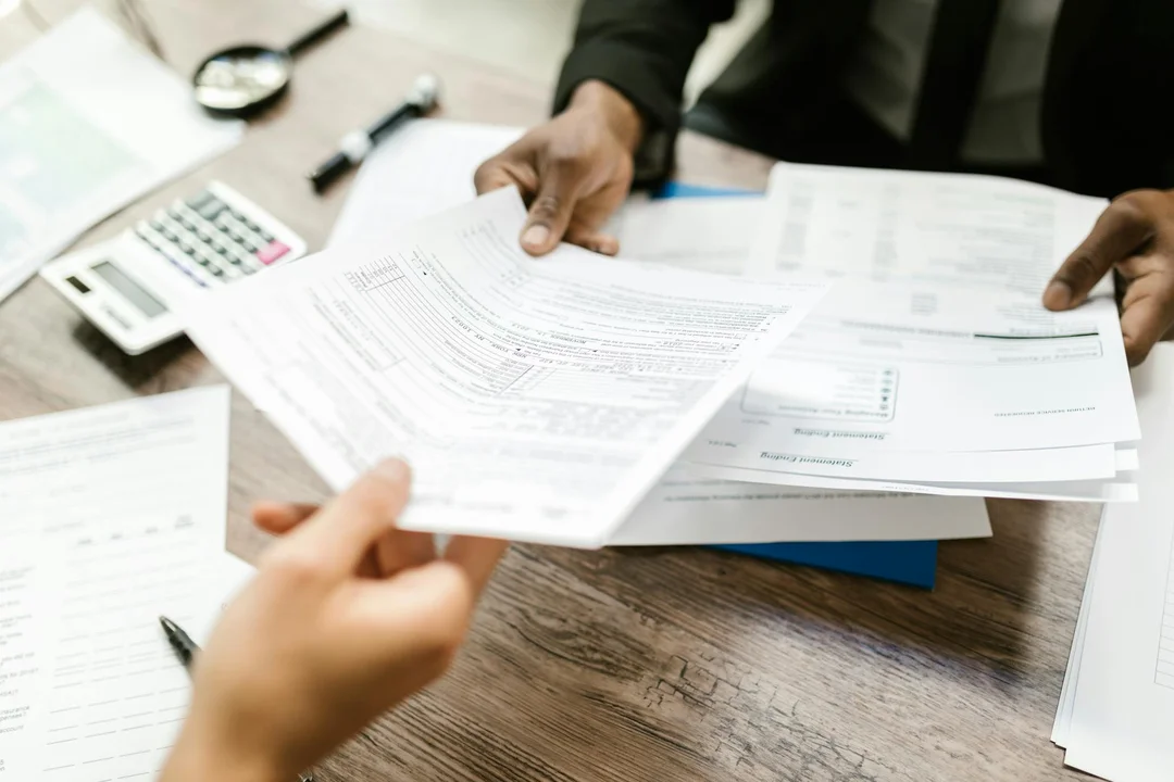 Close-up of hands sorting and reviewing printed meeting documents on a wooden desk, with a calculator and pen nearby.