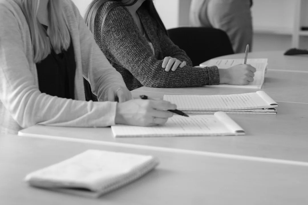 Black-and-white image of several people seated at a table, taking notes and writing in notebooks during a meeting