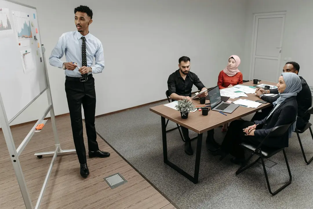 Presenter standing by a whiteboard while a group of people sit around a conference table in a meeting room