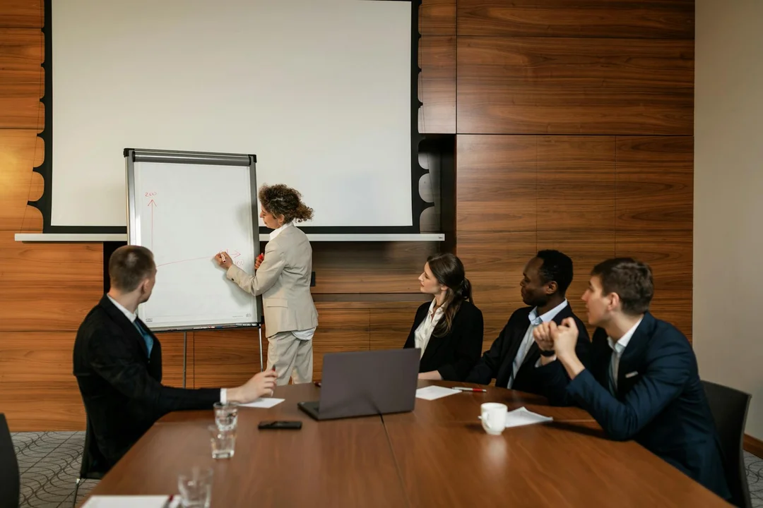 A presenter writes on a whiteboard at the front of a conference room while four attendees sit around a long table, listening and taking notes.