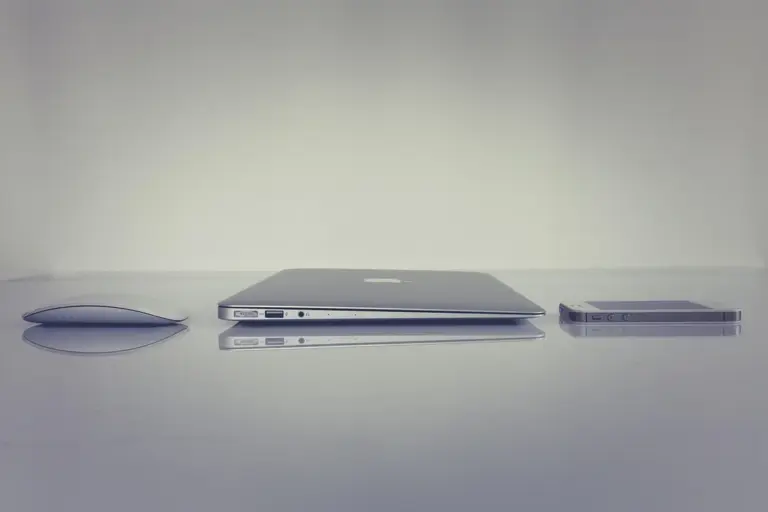 A minimalist desk scene with a closed laptop, a pair of glasses, and a slim device on a white surface