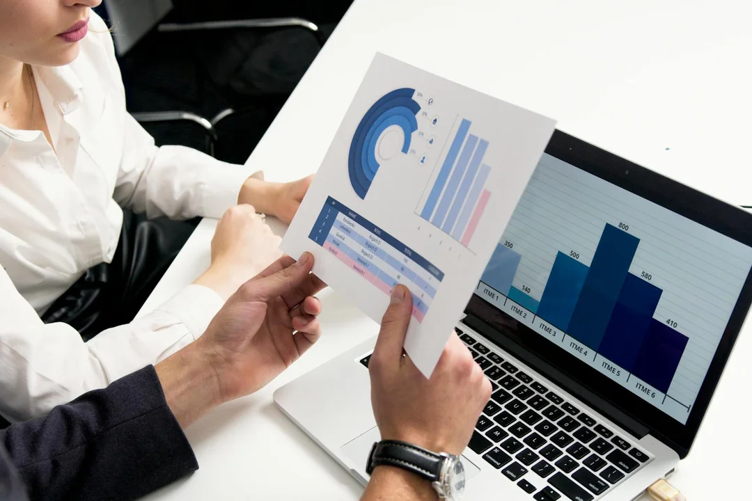 Two professionals review printed charts beside an open laptop displaying graphs, illustrating data analysis during an HOA meeting.