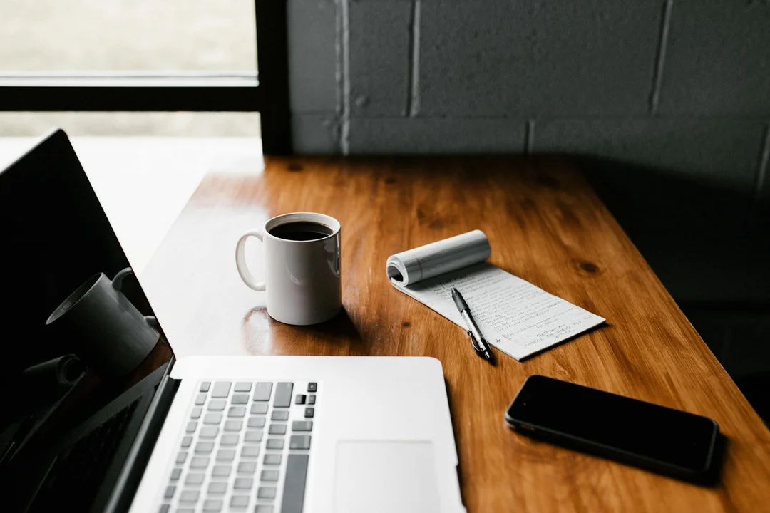 A laptop, coffee mug, notebook, and smartphone on a wooden desk near a window.
