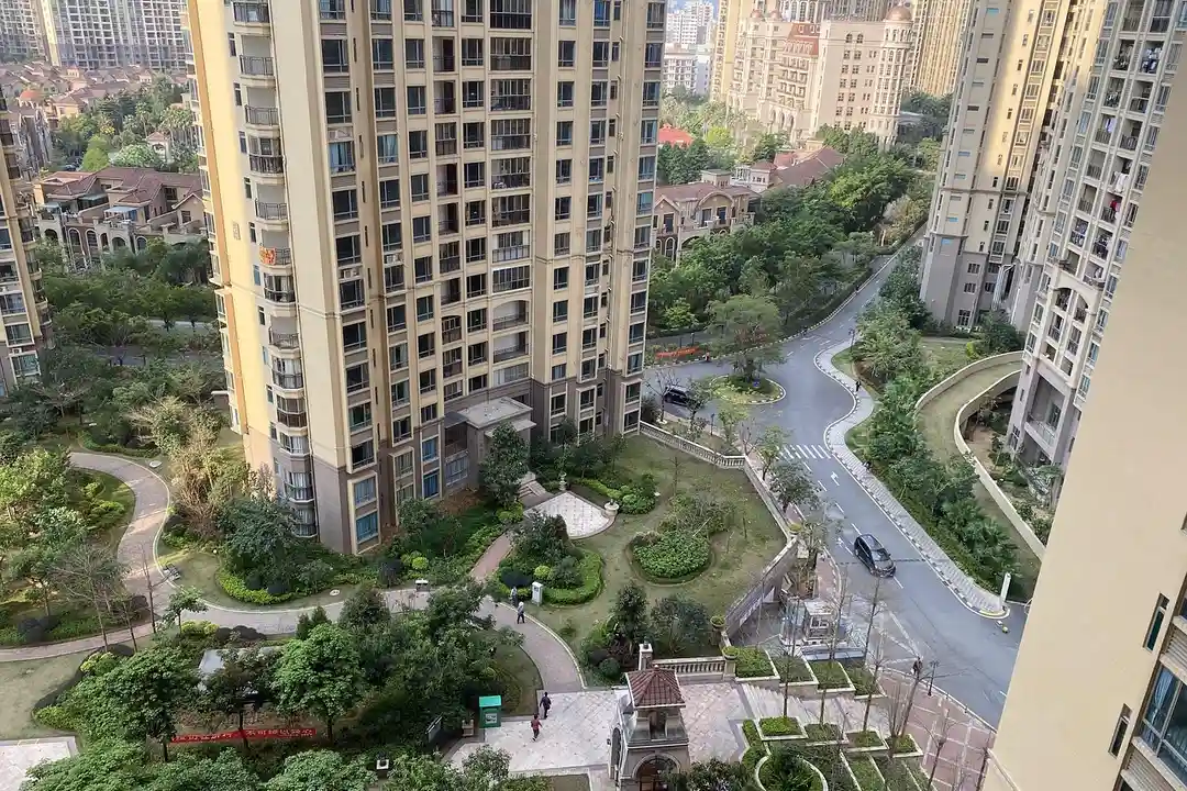 Aerial view of a modern residential complex with tall beige apartment buildings, green courtyards, winding walking paths, and tree-lined roads.