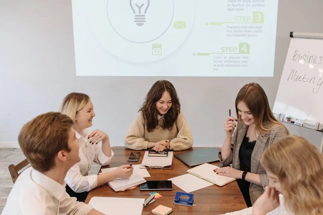 Five professionals around a round table in a meeting room, reviewing documents and taking notes during a discussion.