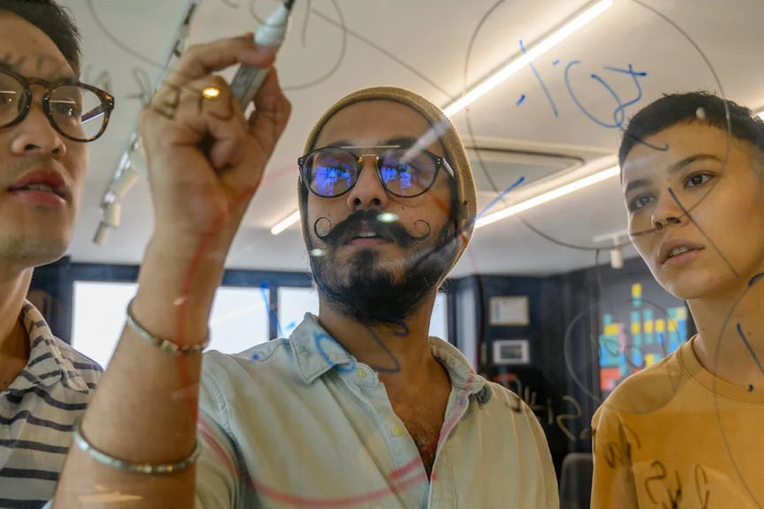 A diverse group of colleagues stands around a glass whiteboard, writing with markers during a collaborative office meeting.