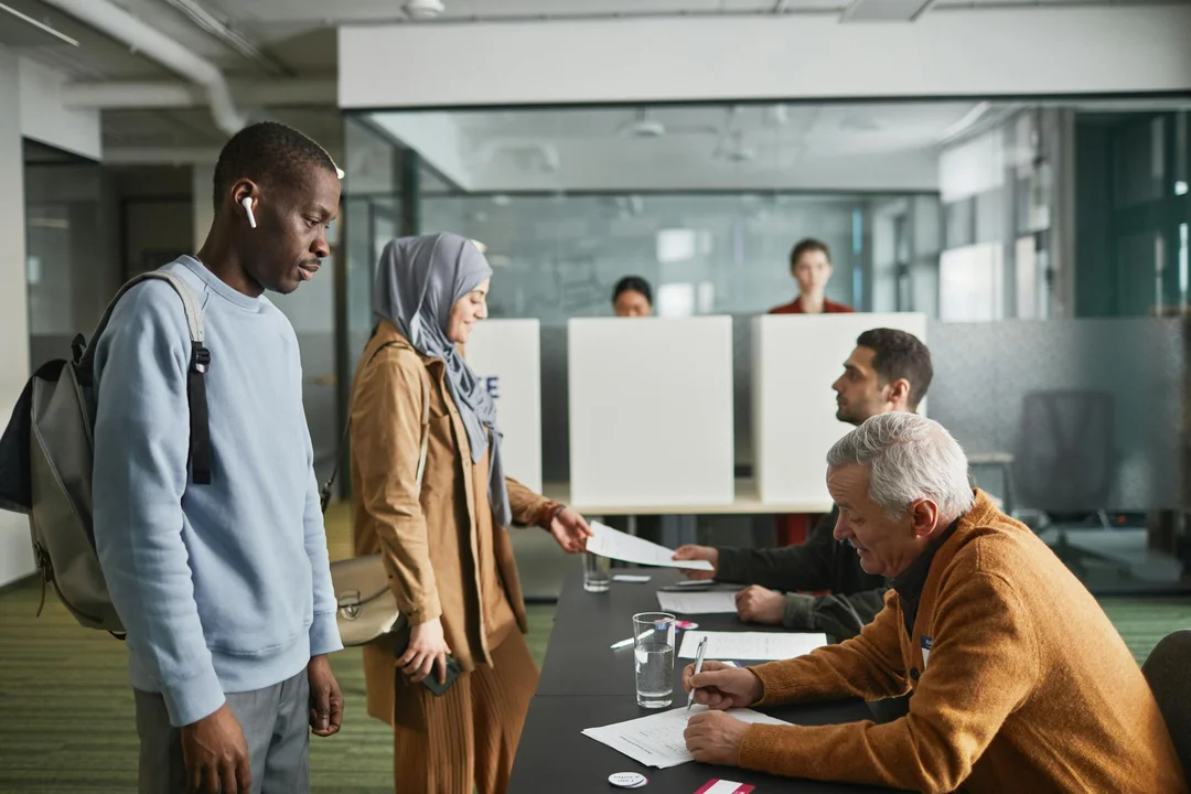 Diverse group of people in an office setting; one person hands documents to a staff member at a desk while others wait.