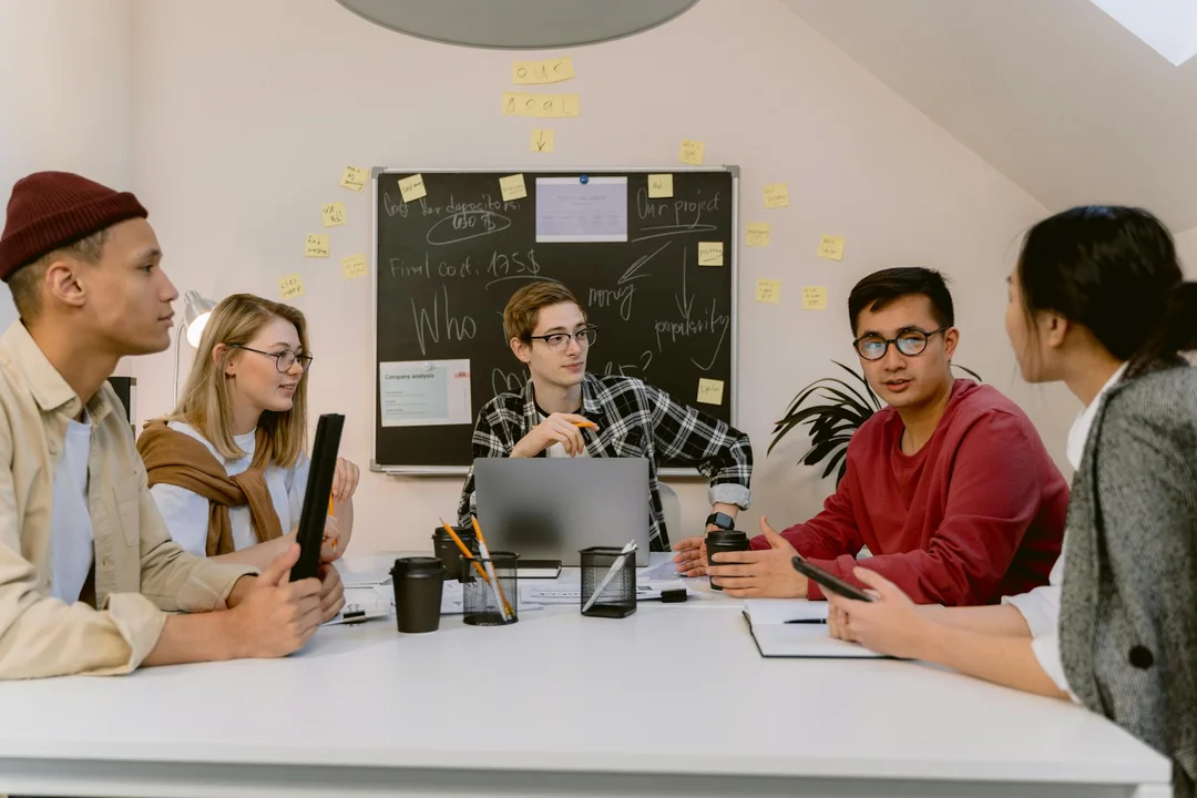 A diverse group of five people seated around a conference table in a modern meeting room, discussing with a laptop and notes, with a sticky-note board in the background.