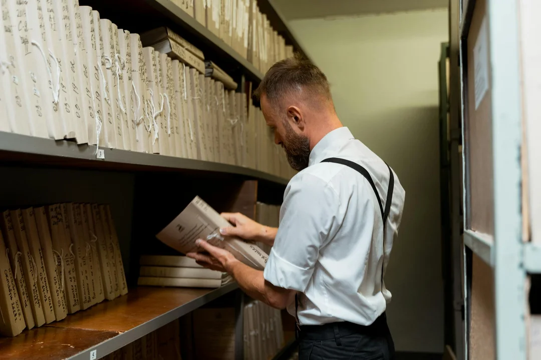 A man in a light blue shirt with suspenders organizes documents on shelves in a records storage room.