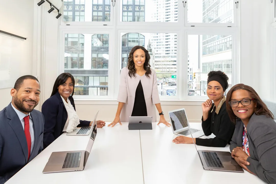 Diverse group of professionals seated around a conference table with laptops, in a bright office with large windows.