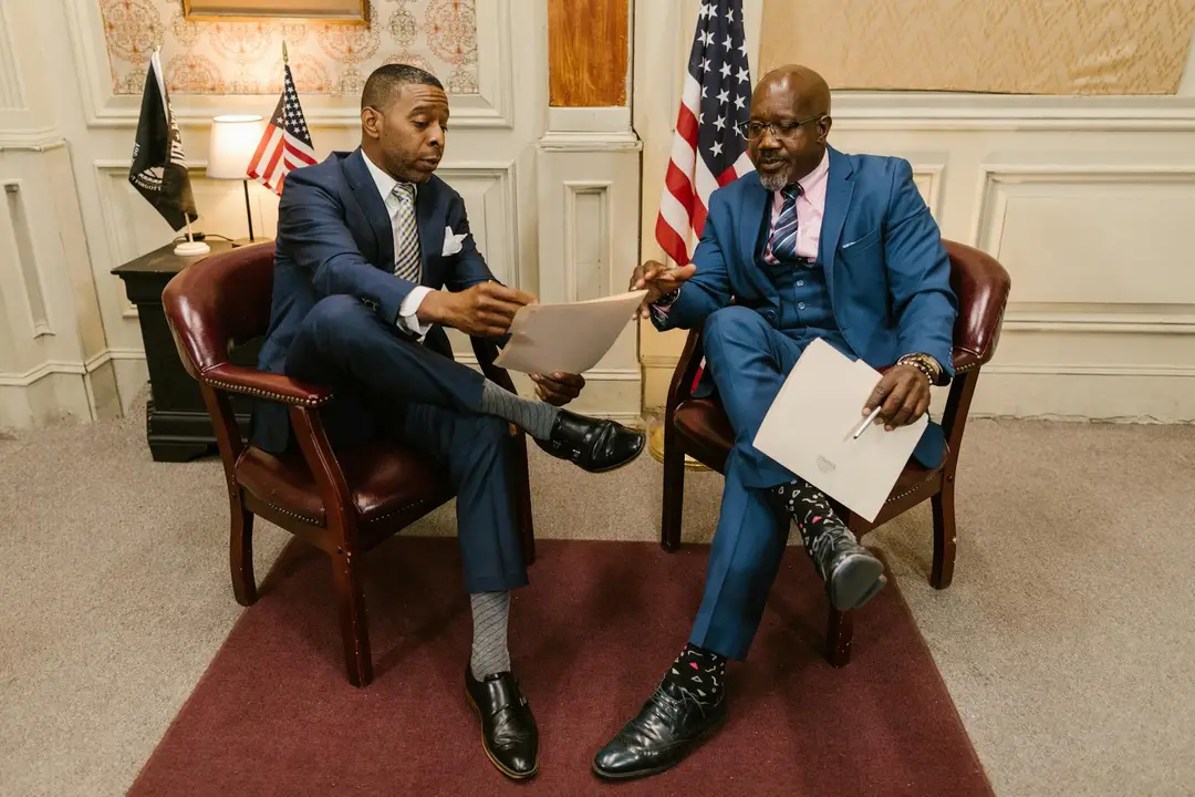Two men in formal business attire sit in leather chairs in a government-like room, reviewing documents as they discuss HOA policy with flags in the background.