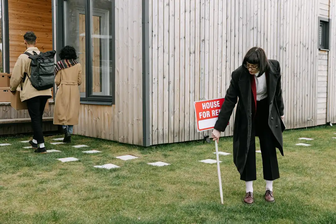 A person holds a 'House for Rent' sign in a grassy HOA community yard while two others walk toward a house in the background.