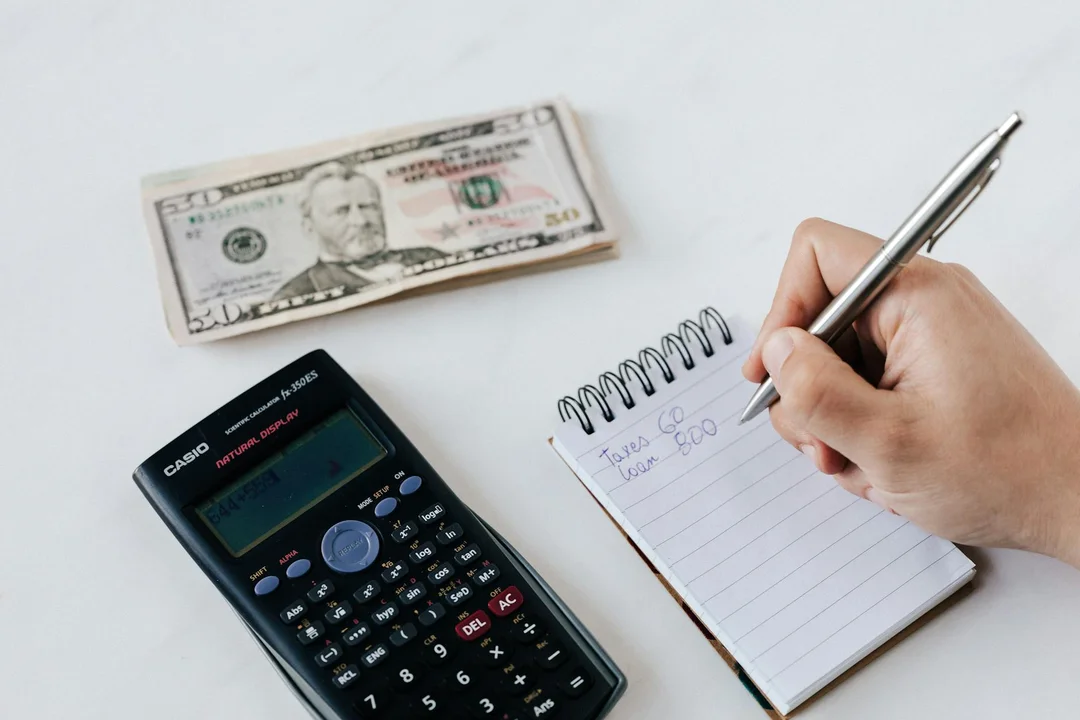 Close-up of a hand writing in a small notebook, with a calculator and a folded dollar bill nearby, symbolizing financial risk and potential liability related to underfunded HOA reserves.