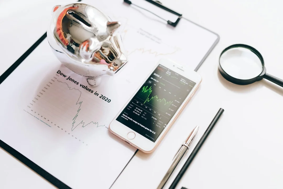 Desk scene with a piggy bank, financial charts on paper, a smartphone displaying market data, a magnifying glass, and a pen, illustrating funding and management of HOA reserve funds.