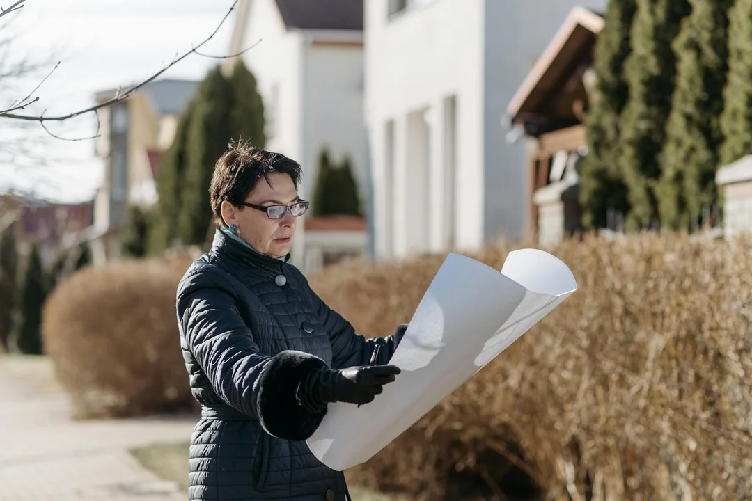 Person wearing a dark jacket and glasses examines a large blueprint outdoors in a suburban neighborhood.
